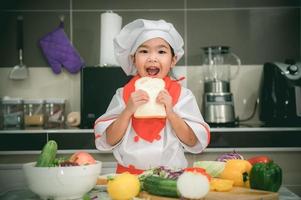 linda asiático niña vestir cocinero uniforme con un lote de vegetal en el mesa en el cocina habitación, hacer comida para comer cena, divertido hora para niños foto