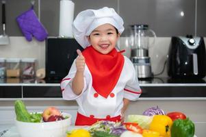 linda asiático niña vestir cocinero uniforme con un lote de vegetal en el mesa en el cocina habitación, hacer comida para comer cena, divertido hora para niños foto