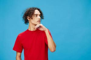 Pensive thoughtful unhappy young curly tanned Caucasian guy in basic red t-shirt posing isolated on over blue background. Lifestyle and Emotions concept. Good offer with copy space for ad photo