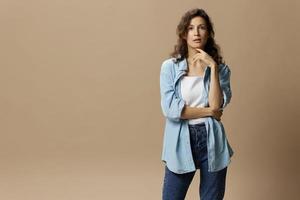 Thoughtful pensive curly beautiful female in jeans casual shirt cross hands recline on hand posing isolated on over beige pastel background. People Lifestyle emotions concept. Copy space photo