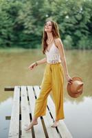 A young woman in a hippie look and eco-dress dancing outdoors by the lake wearing a hat and yellow pants in the summer sunset photo