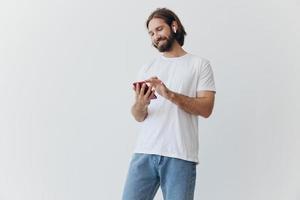 A man with a beard and long hair in a white T-shirt and blue jeans looks at his phone flipping through an online social media feed with headphones in his ears against a white background photo