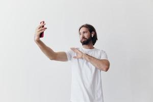 un masculino blogger grabando en video él mismo en su teléfono y chateando con personas en línea con un sonrisa en un blanco camiseta en contra un blanco pared foto