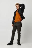 sexy man in a leather jacket holds his hand behind his head and light background orange sweater pants photo