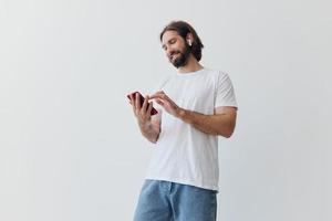 un hombre con un barba y largo pelo en un blanco camiseta y azul pantalones mira a su teléfono voltear mediante un en línea social medios de comunicación alimentar con auriculares en su orejas en contra un blanco antecedentes foto