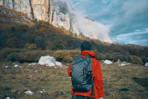 a woman in a red jacket with a backpack and in a warm hat walks on the grass in the fall in the mountains photo