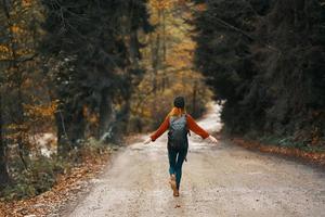 woman in jeans jacket with a backpack walks along the road in the autumn forest photo