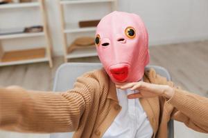 gracioso rubia dama en rosado pescado máscara calentar suéter haciendo selfie utilizar teléfono sentado en Sillón a moderno hogar interior. pausa desde trabajar, tomar un romper, social medios de comunicación en gratis hora concepto. amplio ángulo foto