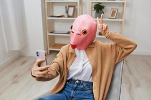 gracioso rubia dama en rosado pescado máscara calentar suéter haciendo selfie utilizar espectáculo v firmar gesto teléfono en Sillón a moderno hogar. pausa desde trabajar, tomar un romper, social medios de comunicación en gratis hora concepto. amplio ángulo foto