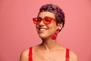 Moda retrato de un mujer con un corto Corte de pelo en de colores Gafas de sol con raro accesorios con pendientes sonriente en un rosado brillante antecedentes foto