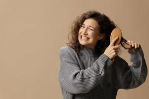 Suffering curly beautiful female in gray casual sweater with hairbrush comb pulls tangled hair posing isolated on over beige pastel background. Problematic unruly damaged hair concept. Copy space photo