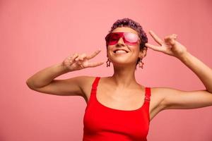 Moda retrato de un mujer con un corto Corte de pelo en de colores Gafas de sol con raro accesorios con pendientes sonrisas en un rosado brillante antecedentes muestra su manos a el cámara foto