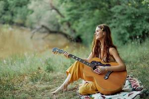 hippie mujer eco simpático jugando guitarra sonrisas y canto canciones en naturaleza sentado en un tartán por el lago en el noche en el puesta de sol luz de sol. un estilo de vida en armonía con el cuerpo y naturaleza foto