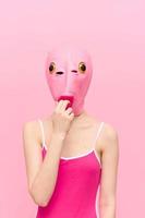 A funny woman wearing a pink fish mask on her head put her fingers in her mouth with pink clothes on a pink background. The concept of modern art photography photo