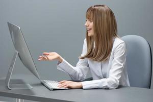 Cheerful pretty blonde businesswoman worker freelancer do video call greeting colleagues using desktop computer in gray modern office. Remote Job, Technology And Career Profession Concept. Copy space photo