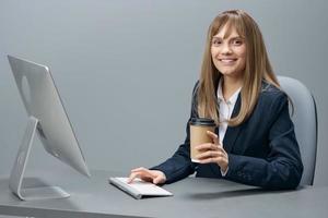 disfruté milenario rubia mujer de negocios trabajador en azul chaqueta utilizando escritorio computadora con para llevar café en gris moderno oficina. remoto trabajo, tecnología y carrera profesión concepto. Copiar espacio foto