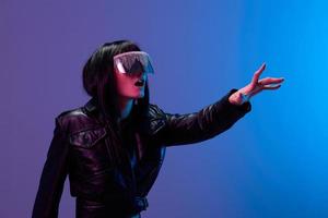 Confused pretty awesome brunet lady in leather jacket trendy specular sunglasses touch invisible object posing isolated in blue violet color light background. Neon party Cyberpunk concept. Copy space photo