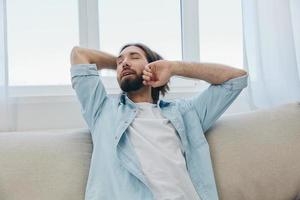 un hombre se sienta en el sofá bostezando y extensión después un siesta, carencia de dormir y fatiga desde trabajo y incorrecto diario rutina. foto