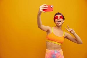 Woman smiles and shows her tongue in unusual glasses millennial takes selfies on her phone in sportswear against an orange studio backdrop, free space photo