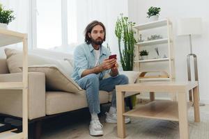 A freelance man in a white T-shirt, blue jeans, and shirt sits on the couch with his phone at home on his day off and plays games photo