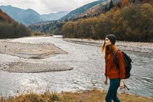 a woman in a red jacket and a backpack on her back are walking on the banks of the river in the mountains photo