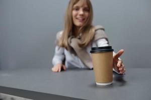 Happy young blonde student lady in warm sweater pulls hand with takeaway coffee at camera sitting at workplace in gray modern home office. Coffee break Lover Concept. Copy space Cool offer Wide Angle photo