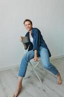 A young woman sitting in a chair at home smiling with teeth with a short haircut in jeans and a denim shirt on a white background. Girl natural poses with no filters photo