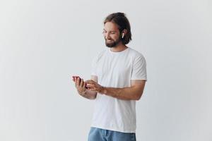 A man with a beard and long hair in a white T-shirt and blue jeans looks at his phone flipping through an online social media feed with headphones in his ears against a white background photo
