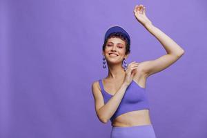 Deportes Moda mujer bailando posando sonriente con dientes en un púrpura Deportes traje para yoga en un esbelto cuerpo y un transparente gorra en su cabeza en un púrpura antecedentes monocromo foto