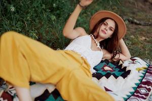 un mujer en Respetuoso del medio ambiente ropa en un hippie Mira es acostado en un de colores tartán sonriente y mirando a un otoño puesta de sol en naturaleza en el parque. estilo de vida en un viaje de armonía con el mundo foto