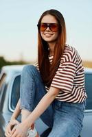 A fashion woman in stylish sunglasses, a striped t-shirt and jeans sits on the trunk of a car and looks at the beautiful nature of autumn. Travel lifestyle photo