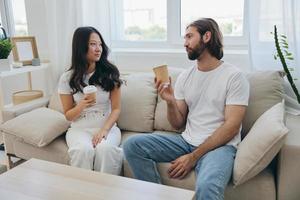 Man and woman sitting at home on the couch in white stylish t-shirts drinking coffee out of crab cups from a coffee shop and having fun chatting smiles and laughter at home. Male and female friendship photo