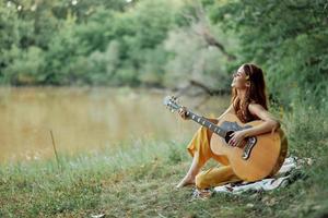 hippie mujer jugando guitarra sonriente y canto canciones en naturaleza sentado en un tartán por el lago en el noche en el rayos de el ajuste Dom. un estilo de vida en armonía con el cuerpo y naturaleza foto