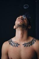 Modelling snapshots. Pensive focused tanned attractive handsome naked man tipping his head back smoking posing isolated in black studio background. Fashion offer. Copy space for ad. Closeup photo