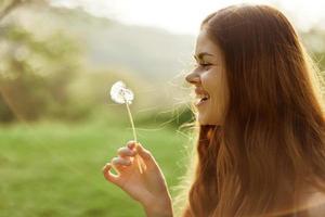 retrato de un joven mujer en perfil con un diente de león flor en su mano soplo en eso y sonriente en contra el verde verano césped en el ajuste Dom en naturaleza foto