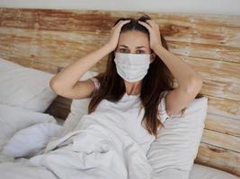 a woman in a medical mask lies in bed under the covers and touches her head with her hands photo