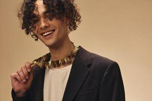 Flirting cute awesome tanned curly man in classic brown jacket ethnic necklace posing isolated on over beige pastel background. Fashion New Collection offer. Retro style concept. Free place for ad photo