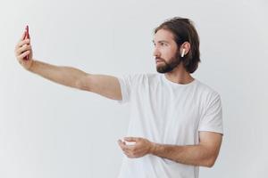 A man with a beard blogger in a white T-shirt with a phone and wireless headphones talking on an online video call against a white background photo