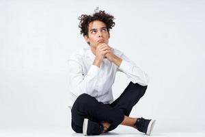 guy with curly hair in a white shirt, sneakers and trousers sits on the floor photo