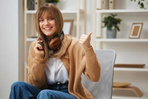 Cheerful lovely young blonde student lady in warm sweater in headphones point thumb up gesture Like sitting in armchair at modern home interior. Music time Relaxing Cool playlist Concept. Cool offer photo