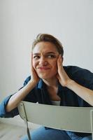 A young woman sitting in a chair at home smiling with teeth with a short haircut in jeans and a denim shirt on a white background. Girl natural poses with no filters photo