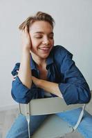 A young woman sitting in a chair at home smiling with teeth with a short haircut in jeans and a denim shirt on a white background. Girl natural poses with no filters photo