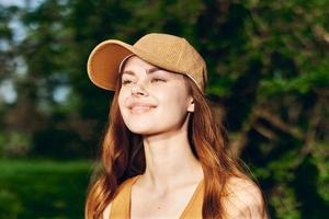 mujer blogger con teléfono en mano en naturaleza en contra un fondo de verdor sonriente en el Brillo Solar vistiendo un gorra después hacer ejercicio foto