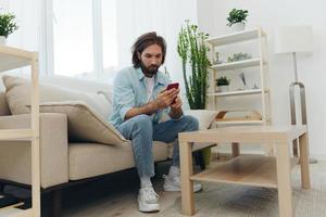 A freelance man in a white T-shirt, blue jeans, and shirt sits on the couch with his phone at home on his day off and plays games photo