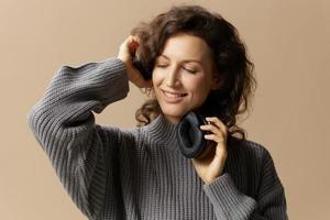 Happy cheerful curly beautiful female in gray casual sweater touched headphones smiling closing eyes posing isolated on over beige pastel background. Sound streaming platform concept. Copy space photo