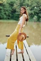 A young woman in a hippie look and eco-dress dancing outdoors by the lake wearing a hat and yellow pants in the summer sunset photo