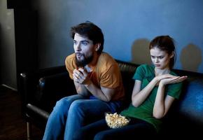 A woman and a man with popcorn on a leather sofa indoors watching TV in the evening photo