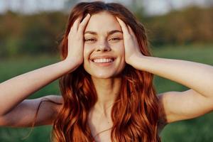A beautiful woman with long, wavy red hair smiles for the camera in nature illuminated by sunset sunlight. The concept of beauty and harmony with nature photo