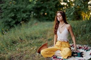 A young hippie woman meditates in nature in the park, sitting in a lotus position on her colorful plaid and enjoying harmony with the world in eco-clothing photo