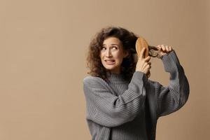 Tormented curly beautiful female in gray casual sweater with hairbrush comb pulls tangled hair posing isolated on over beige pastel background. Problematic unruly damaged hair concept. Copy space photo